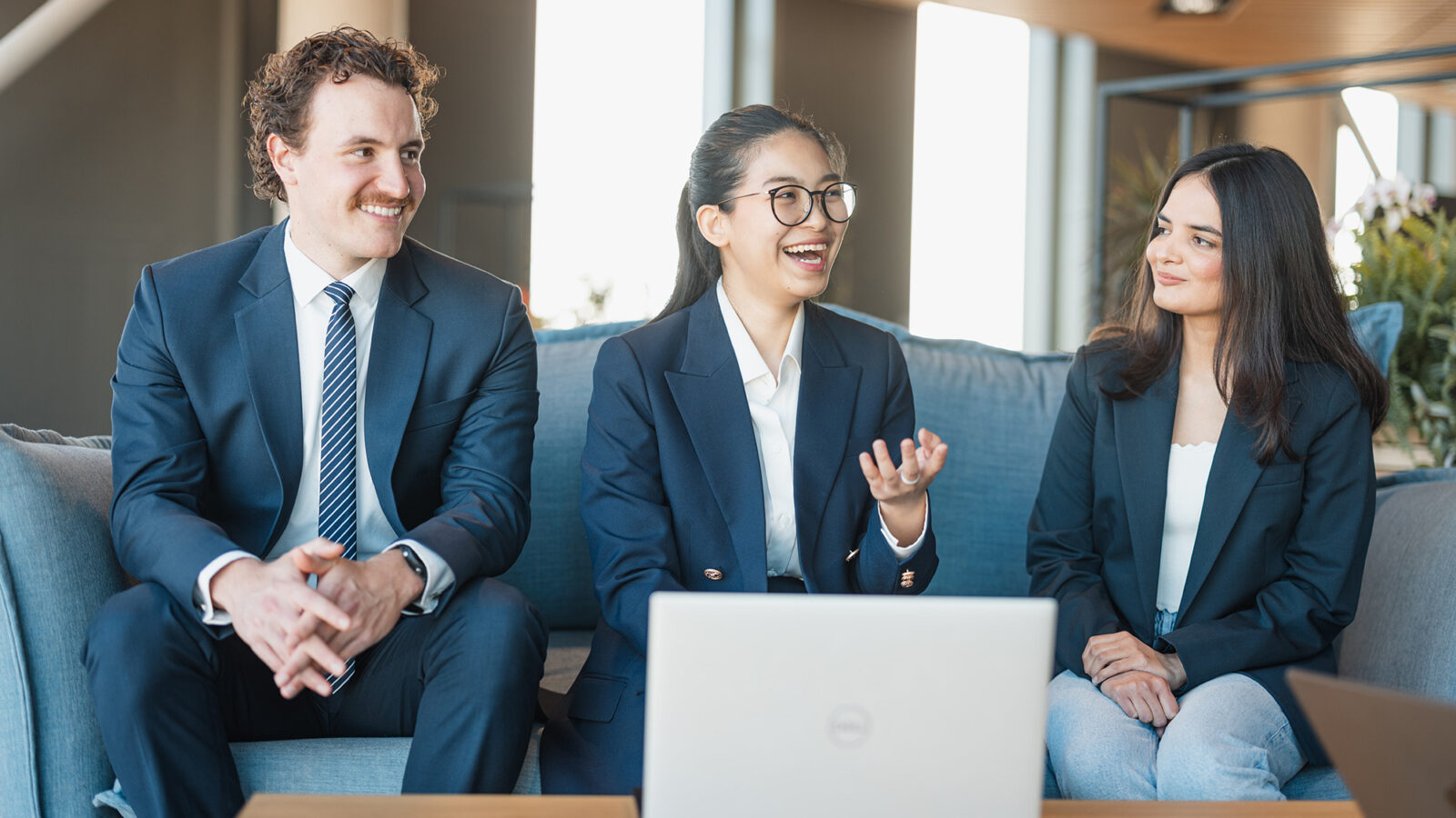Three students in suits sitting round a laptop smiling
